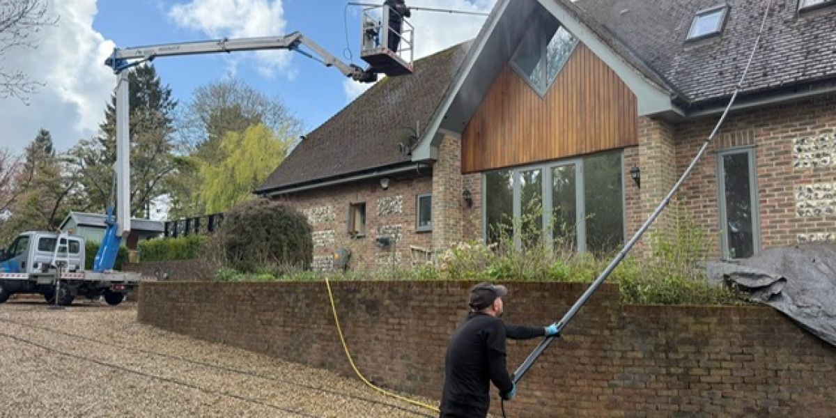 A man is cleaning the roof of a house using a broom and ladder under a clear blue sky.