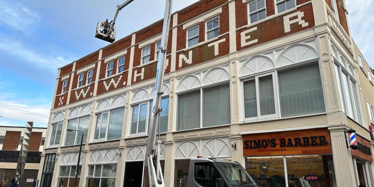 Worker in a truck-mounted cherry picker accessing the roofline of a brick building above Simo’s Barber shop.