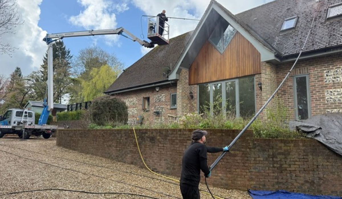 A man is cleaning the roof of a house using a broom and ladder under a clear blue sky.